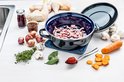 Zwarte geëmailleerde pan met rauwe spekblokjes, omringd door ingrediënten zoals kip, uien en wortelen.
