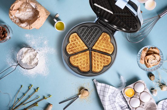 Blauw wafelijzer met vijf hartvormige wafels, omringd door bakbenodigdheden zoals bloem, eieren en fruit.