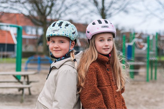 Twee meisjes op een speelplaats. Een draagt een lichtgroene helm met winter/dierenmotief, de ander een roze helm.