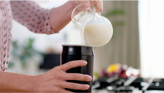 Een persoon schenkt melk uit een glazen karaf in een zwarte melkopschuimer.