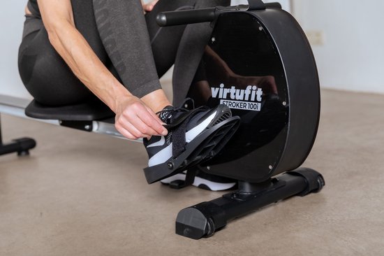 Een vrouw met donkere sportkleding maakt haar schoen vast aan de pedal van de Virtufit roeimachine.