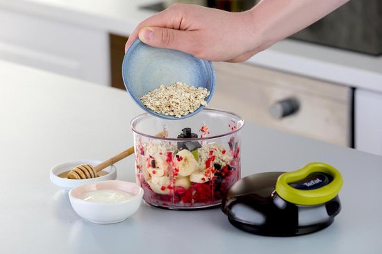 Een hand die havervlokken in een Tefal hakmolen met bananen, bessen en yoghurt doet.