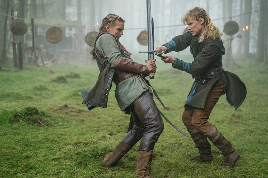 Twee mannen met lang blond haar, gekleed in historische kleding, oefenen met zwaarden in een bosrijke omgeving.