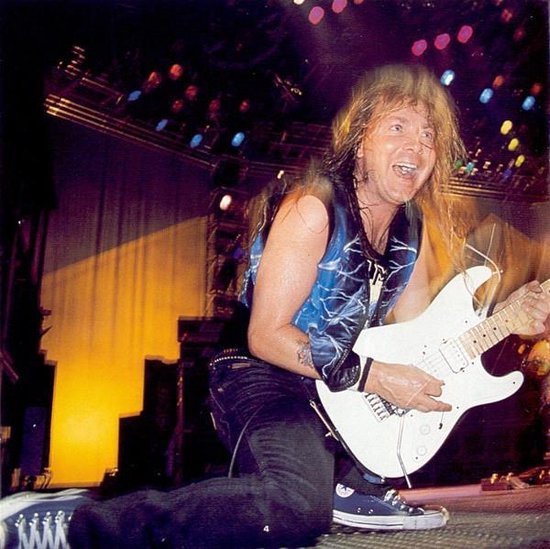 Een gitarist met lang blond haar speelt op een witte elektrische gitaar, knielend op een podium met felle lichten.