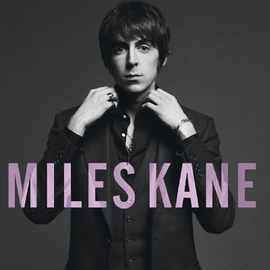 Miles Kane in zwart pak met vest, donkere blouse en ketting. Hij strijkt zijn kraag glad. Tekst: MILES KANE.