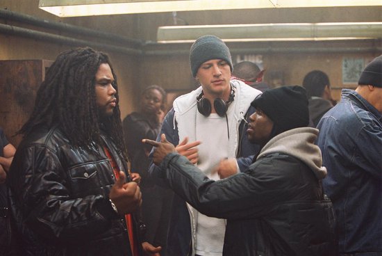 Drie mannen, waaronder een met dreadlocks, praten in een industriële ruimte. Twee dragen mutsen.