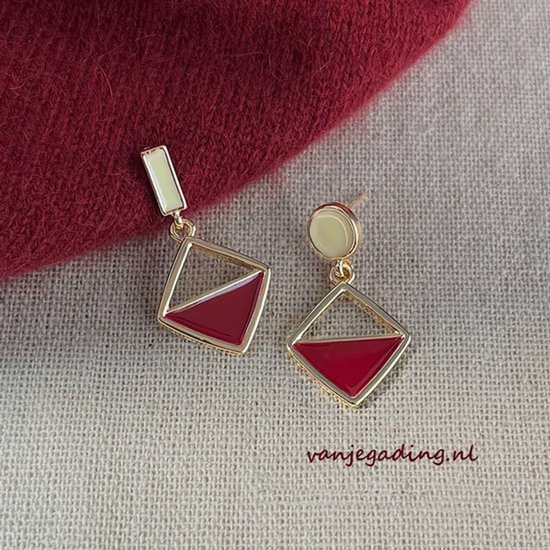 Gouden oorbellen met een rood en geel vierkantje. De oorbellen hangen aan een rode stof.