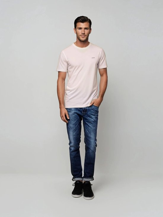 Man met bruin haar draagt een lichtroze t-shirt met opdruk en een spijkerbroek.