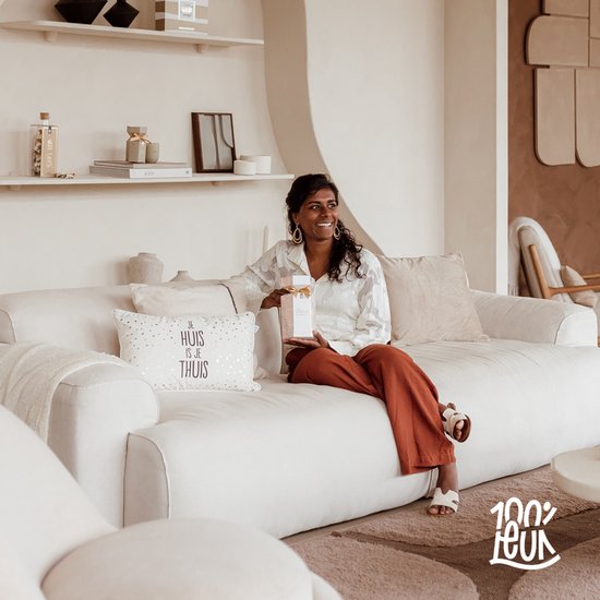 Vrouw in woonkamer op bank met cadeaupakket, houdt product vast. Tekst op kussen 'Je huis is je thuis'.