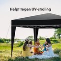 Vrouwen picknicken onder een zwarte partytent in een grasrijk gebied. De tekst "Helpt tegen UV-straling" is zichtbaar.