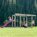 Groep kinderen spelen op een uitgebreid houten speeltoestel met paarse glijbaan en schommels.