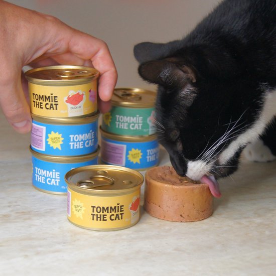Een zwart-witte kat likt kattenvoer uit een open blikje, met stapels blikjes op de achtergrond.