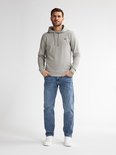 Man in een lichtgrijze hoodie en lichtblauwe jeans, staand met handen in de zakken en witte sneakers.