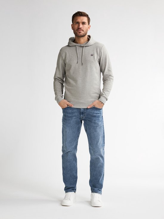 Man in een lichtgrijze hoodie en lichtblauwe jeans, staand met handen in de zakken en witte sneakers.