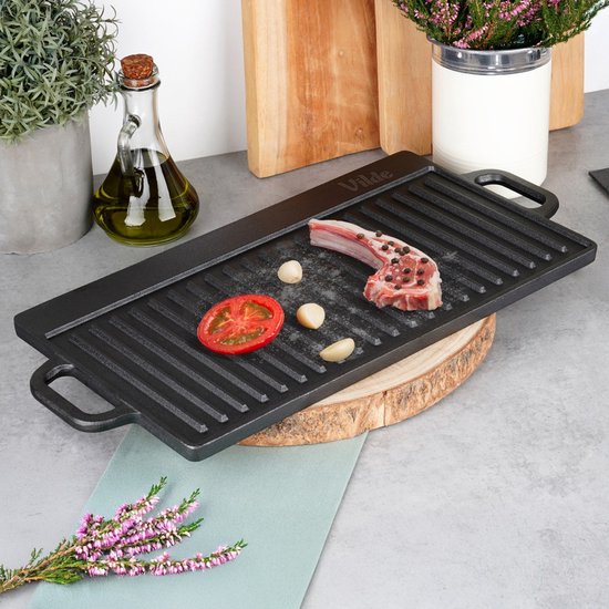 Zwarte gietijzeren grillplaat met ribbels, met een stuk vlees, knoflook en tomaat.