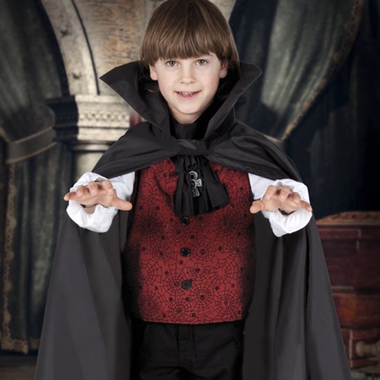 Jongen in vampierkostuum met zwarte cape, rood vest met spinnenwebben, wit shirt en zwarte broek, handen geheven.