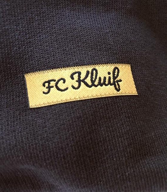 Détail d'un pull foncé avec une étiquette appliquée "FC Kluif" en lettres jaunes.