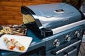 Close-up van een antracietkleurige gasbarbecue met gegrilde groenten en spiesjes op een bord ernaast.