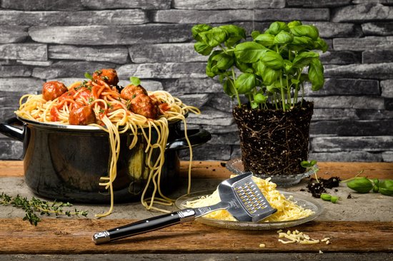 Spaghetti met gehaktballetjes in tomatensaus in een pan, geserveerd met een pot basilicum en een kaasrasp.