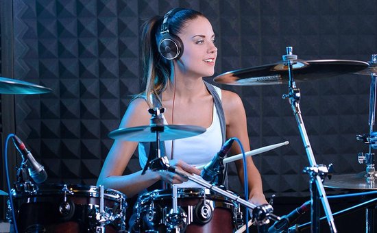 Vrouw met bruine paardenstaart, draagt een witte top en zwarte Studio DJ koptelefoon tijdens het drummen.