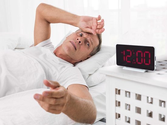 Een man wordt wakker in bed, kijkt verbaasd naar de witte digitale wekker met rood display dat 12:00 toont.