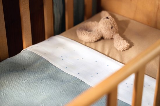 Een babywieg met een bruine teddybeer en een wiegdeken met wit bovenstuk en grijs gebreid onderstuk.