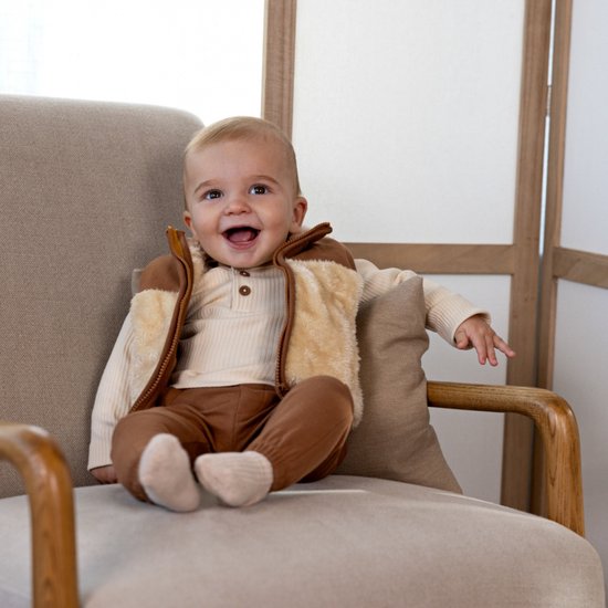 Vrolijke baby in een beige ribtop, een bruine teddy bodywarmer en een bruine joggingbroek, zittend in een fauteuil.