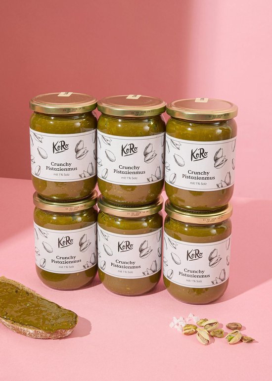 Zes potten KoRo crunchy pistachenotenpasta met 1% zout, op een roze achtergrond met een stuk brood en pistachenoten.