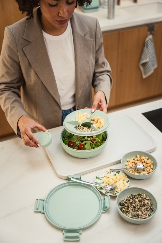 Femme en blazer marron ajoutant des ingrédients sur une boîte à salade vert pâle avec diviseur.