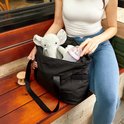 Femme en jean et haut blanc sortant un éléphant en peluche et un biberon d'un sac fourre-tout noir.
