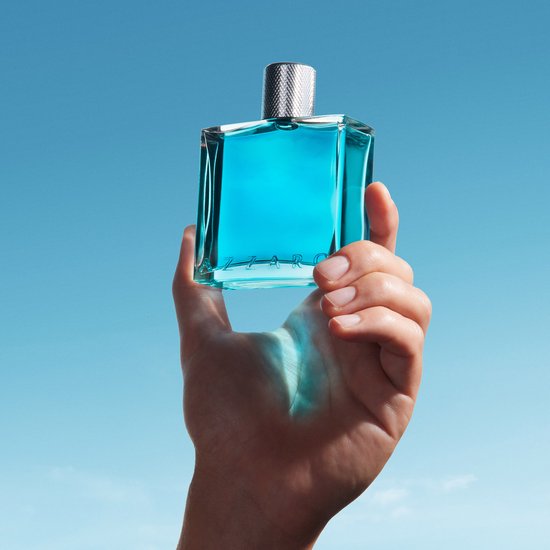 Een hand houdt een helderblauwe parfumfles vast tegen een heldere blauwe hemel.