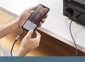 Een hand houdt een smartphone vast met een muziekapp, verbonden via een zwarte adapter met UGREEN-logo met een audio-installatie.