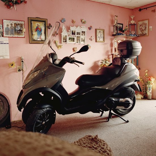 Grijze driewielscooter met zwarte zitting, geparkeerd in een kamer met roze muren en decoraties.