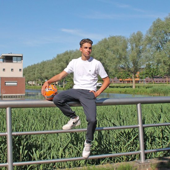 Man zit op een hek met een bal, draagt een donkere joggingbroek en witte t-shirt.