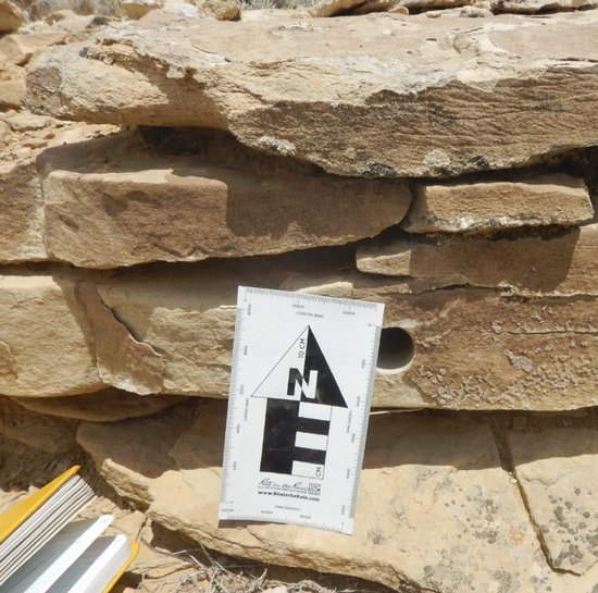 Close-up van gelaagde rotsen met een geologische meetlat in het midden. Een deel van een geel notitieboek is zichtbaar.
