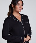 Model met donker haar in een staart draagt een zwart pyjamashirt met witte biezen en opgestikte borstzak.