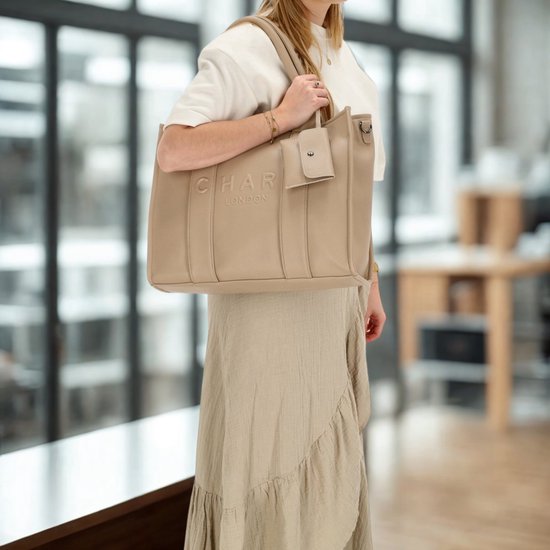 Vrouw met beige handtas met 'CHARM LONDON' logo, draagt deze crossbody met schouderband.