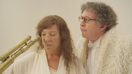 Een vrouw met lang bruin haar en een witte jurk kijkt naar een antieke telescoop, naast een man met grijs krullend haar en bril.
