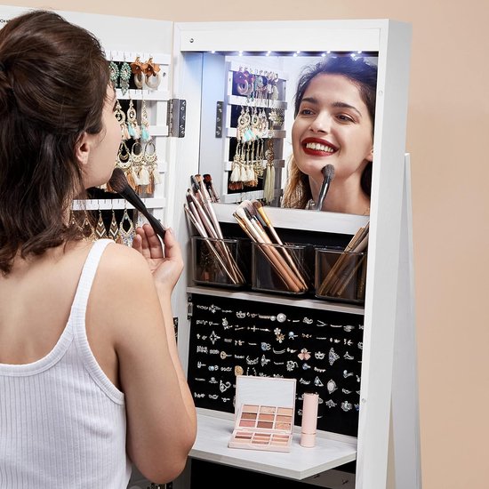 Vrouw met make-up kwast kijkt in witte spiegelkast. Sieraden en make-up zichtbaar.