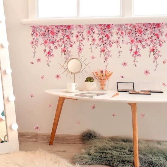 Hangende roze bloesemtakken boven een witte tafel met make-up en accessoires, op een muur met losse bloesems.