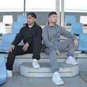 Twee mannen zitten op een trap. De man links draagt een zwarte joggingbroek en hoodie. De man rechts draagt een grijze joggingbroek en hoodie.
