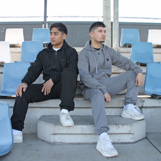 Twee mannen zitten op een trap. De man links draagt een zwarte joggingbroek en hoodie. De man rechts draagt een grijze joggingbroek en hoodie.