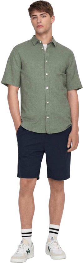 Een man in een lichtgroen linnenmix overhemd met korte mouwen en donkerblauwe shorts.