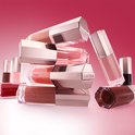 Stapel Lancôme lipgloss flesjes in verschillende roze en bruine tinten, tegen een rode achtergrond.