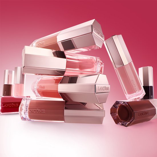 Stapel Lancôme lipgloss flesjes in verschillende roze en bruine tinten, tegen een rode achtergrond.