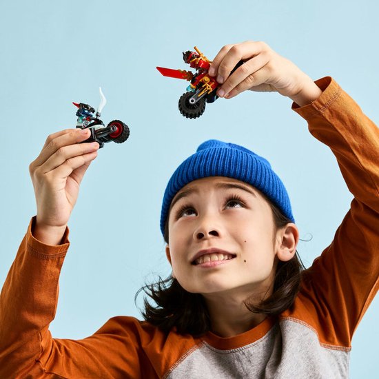 Kind met blauwe muts houdt twee LEGO Ninjago speelgoedmotoren omhoog, één rode en één blauwe.