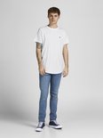 Man in wit t-shirt met kleine zwarte logoprint en jeans, staand.