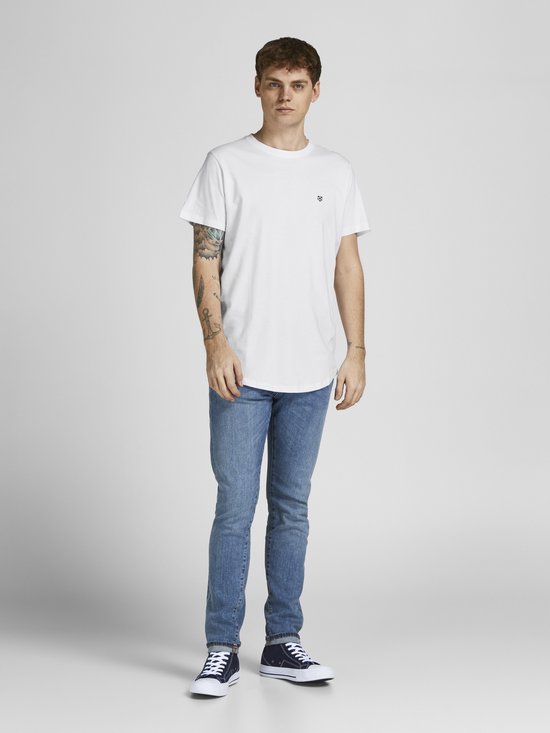 Man in wit t-shirt met kleine zwarte logoprint en jeans, staand.
