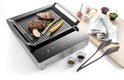 Inductie grillplaat met ribbelgrill, waarop twee steaks, asperges, wortel en knoflook liggen. Naast de grill staan peper- en zoutvaatjes, tang en kruiden.