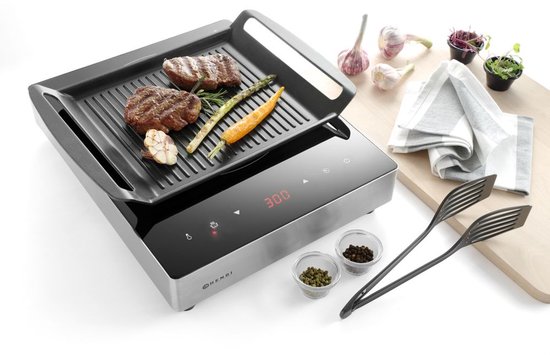 Inductie grillplaat met ribbelgrill, waarop twee steaks, asperges, wortel en knoflook liggen. Naast de grill staan peper- en zoutvaatjes, tang en kruiden.